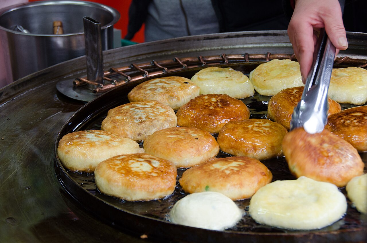 油を引いた鉄板で、ホットクに焼き色がついていきます。トングのそばには、生地を平たく押す丸い道具が置かれています。2013年の資料写真です。