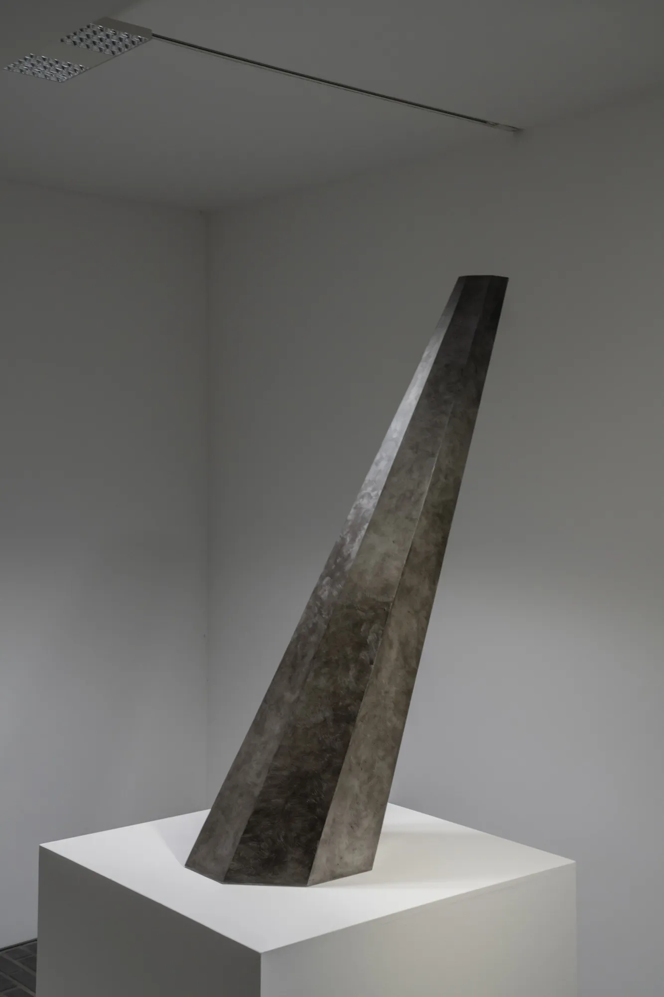 A gray tapered polyhedral sculpture rising at an angle from a white pedestal