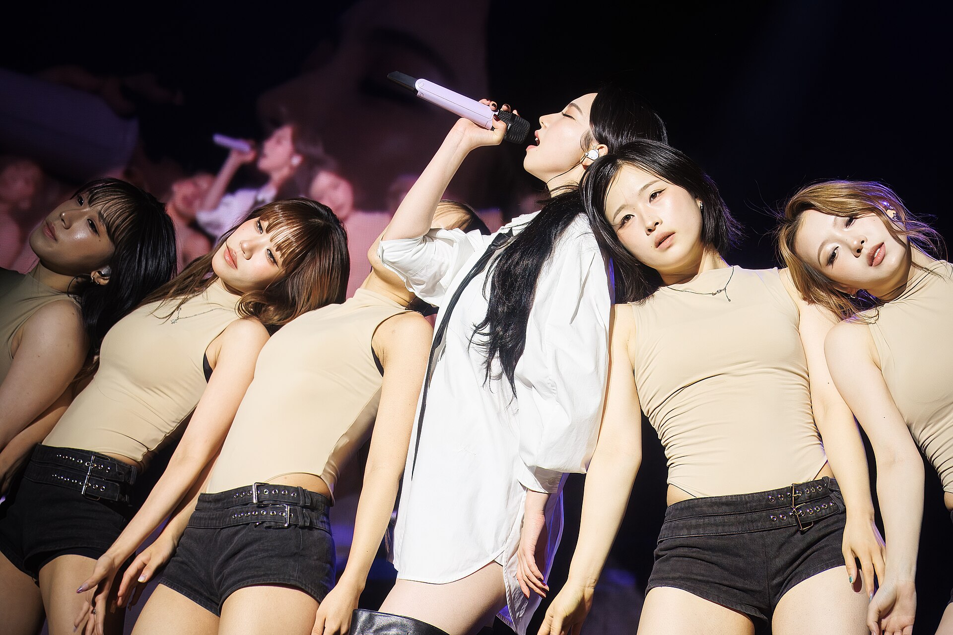 A member in a white shirt singing into a microphone, surrounded by dancers in beige