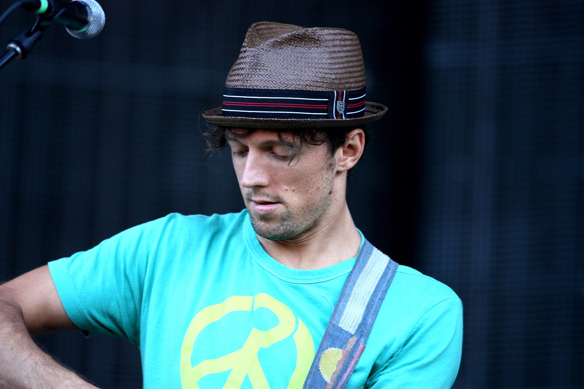 Jason Mraz in a fedora looking down, guitar strap over his shoulder