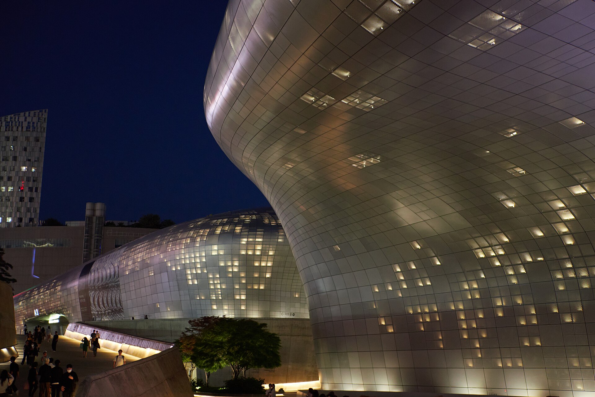The curved aluminium-panel exterior of DDP glowing with window light, people walking below