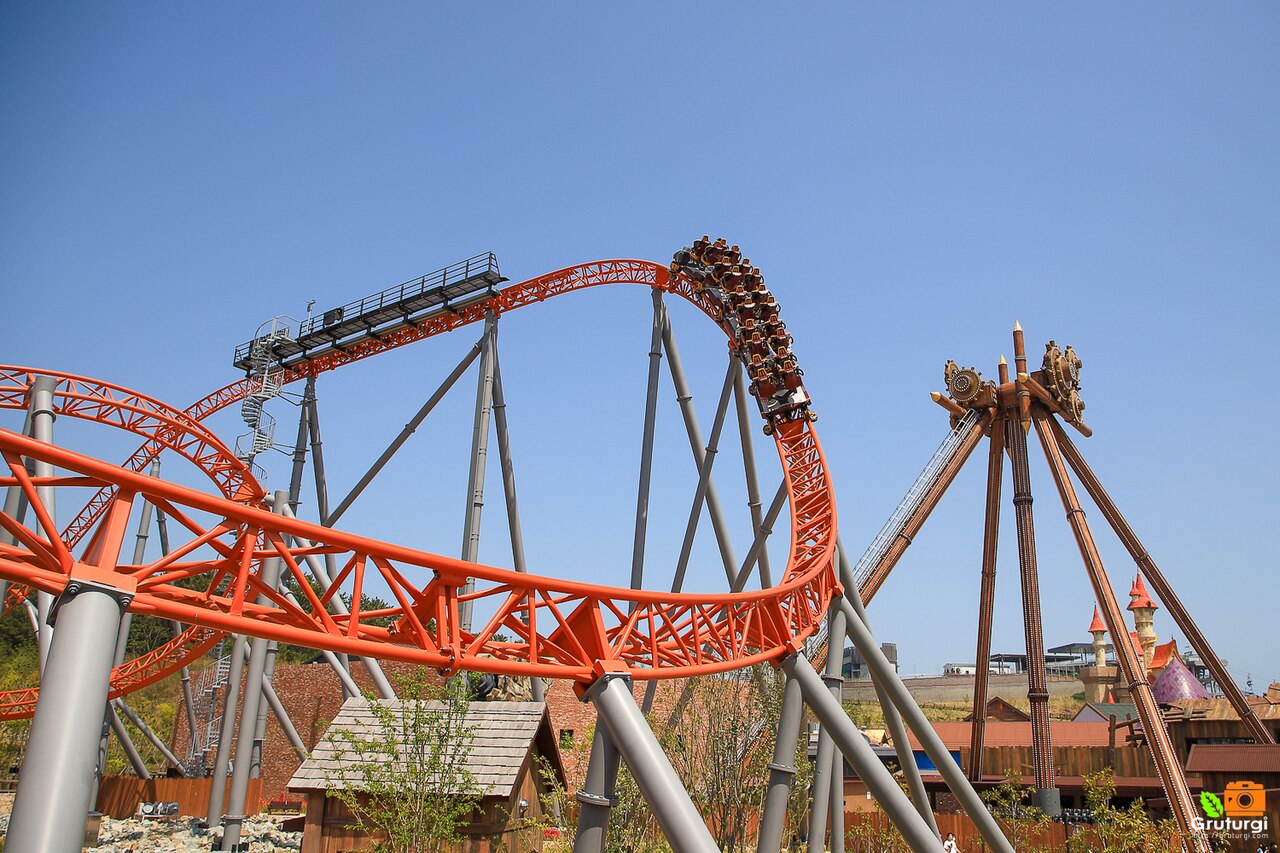 An orange roller coaster track and train against a blue sky, with a brown steel ride on the right