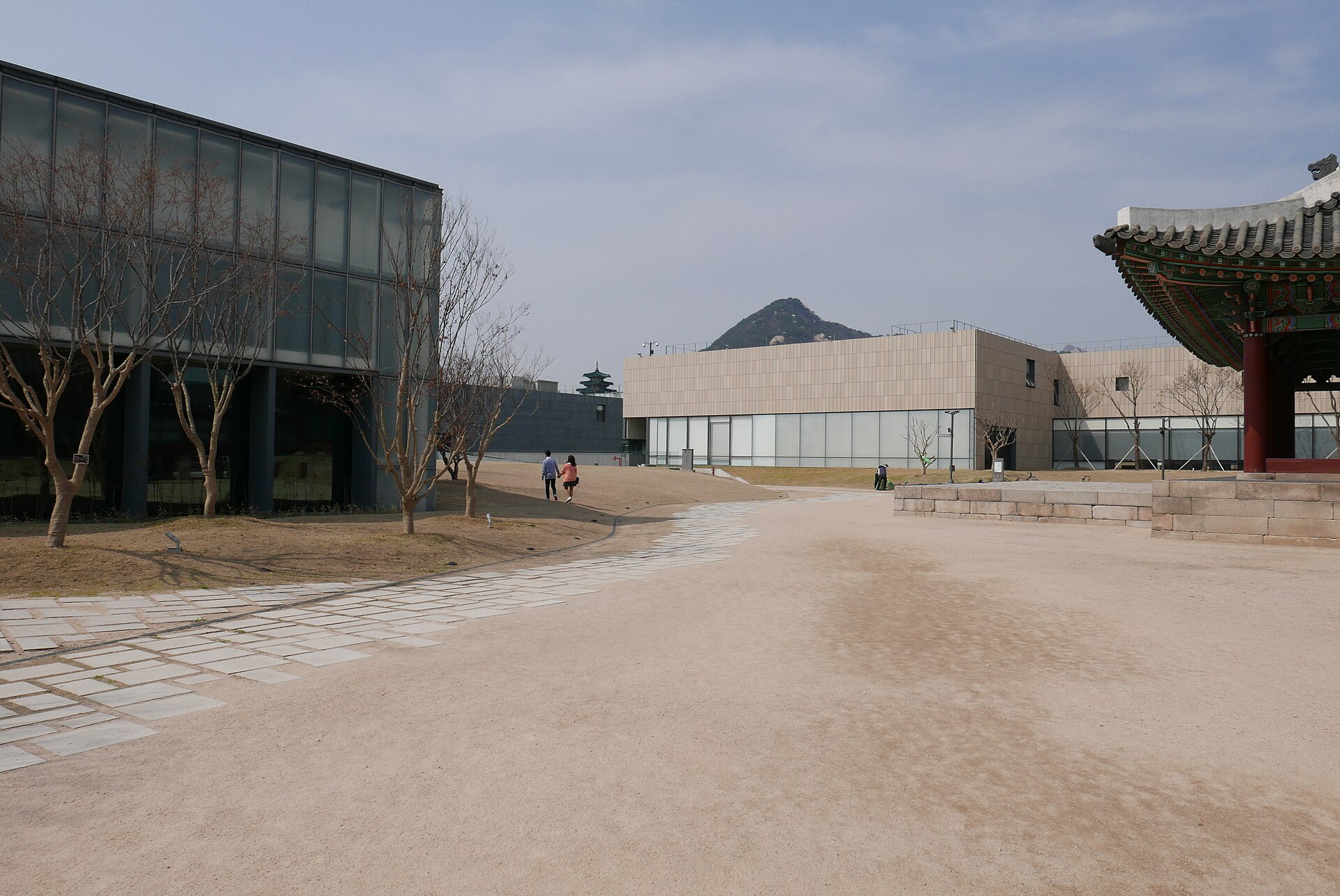 A wide courtyard between a glass building and a hanok roof edge, with a hill beyond