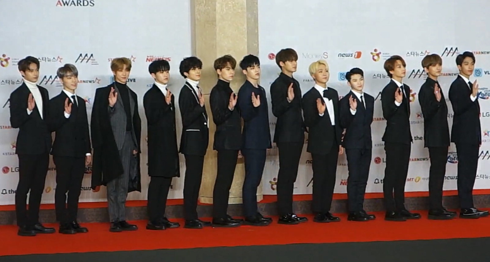 검은 정장을 입은 세븐틴 멤버 13명이 레드카펫 위에 한 줄로 서서 손을 흔드는 모습