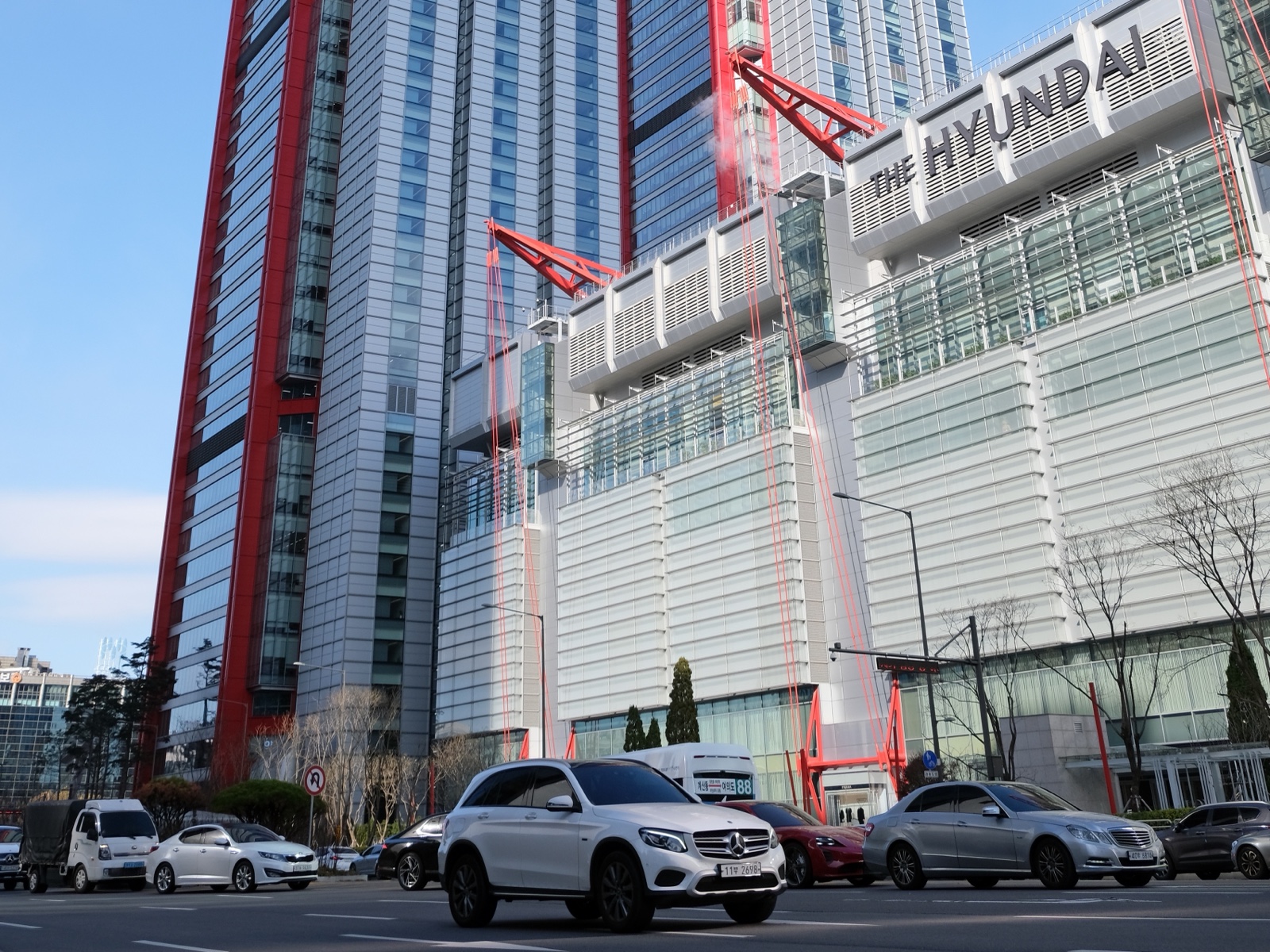 THE HYUNDAI 간판이 달린 흰 건물과 붉은 철골 고층 빌딩, 앞 도로의 자동차들