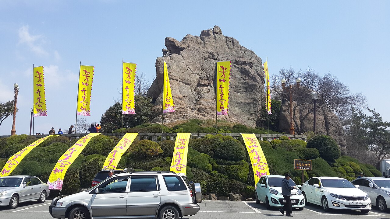 둥근 노적봉 바위 아래에 초목과 노란 봄축제 깃발, 주차된 차들이 있는 풍경